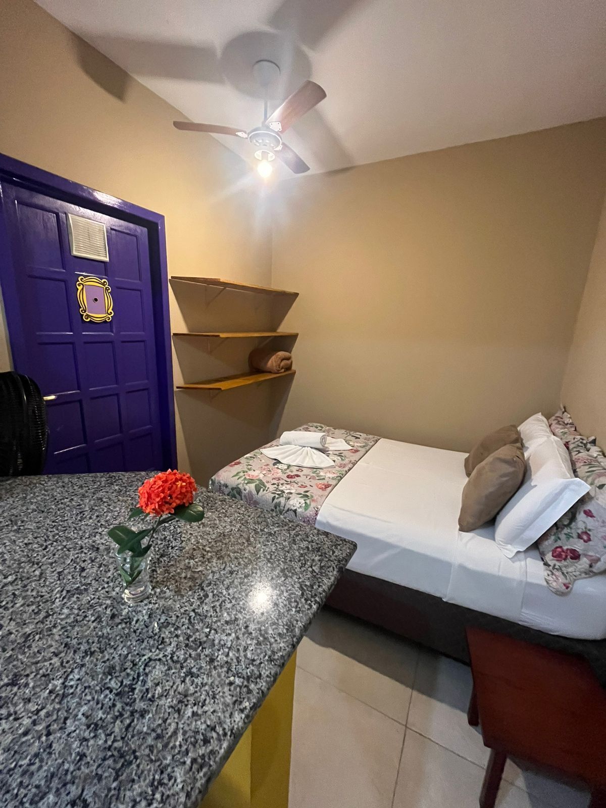 Una habitación con cama, una mesa con una flor, un ventilador de techo y una puerta morada.