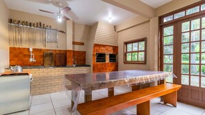 A kitchen with a brick backsplash, a wooden table with a bench, a ceiling fan, and a window with a view of greenery. - Pousada ChaDay (Imbituba)
