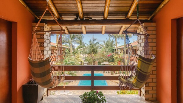 A wooden deck with two hammocks, a potted plant, and a view of a swimming pool and palm trees. - Pousada ChaDay (Imbituba)