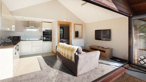 Eine moderne Küche mit weißen Schränken, einer Mikrowelle und einem Herd. Es gibt ein graues Sofa mit einer Decke, eine Holzbank und einen Fernseher an der Wand.