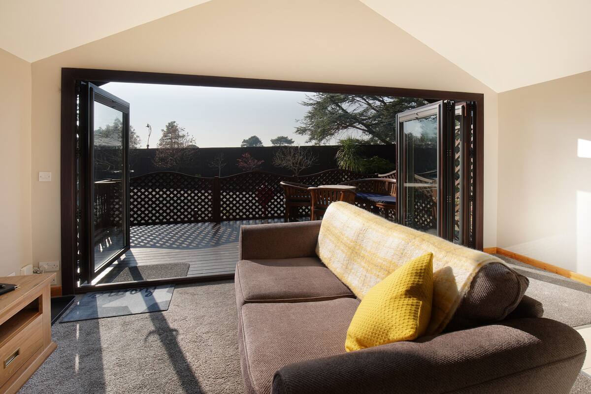 Un salon moderne avec un canapé marron, un coussin jaune et une porte vitrée donnant sur une terrasse extérieure.