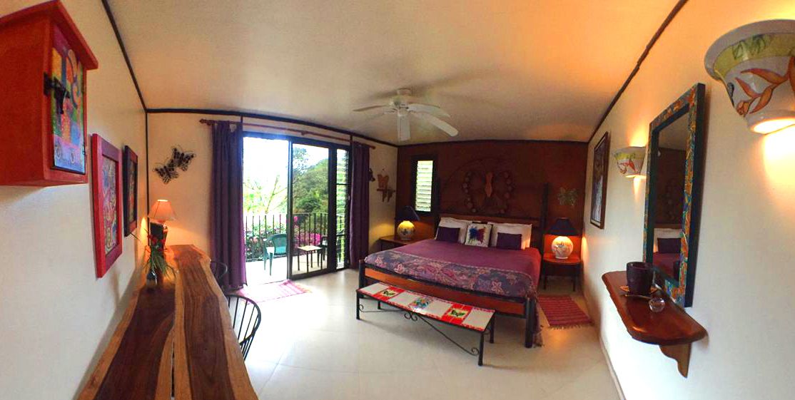Ein Schlafzimmer mit Bett, Schreibtisch, Stuhl, Deckenventilator und einem Balkon mit Blick auf Grünflächen.