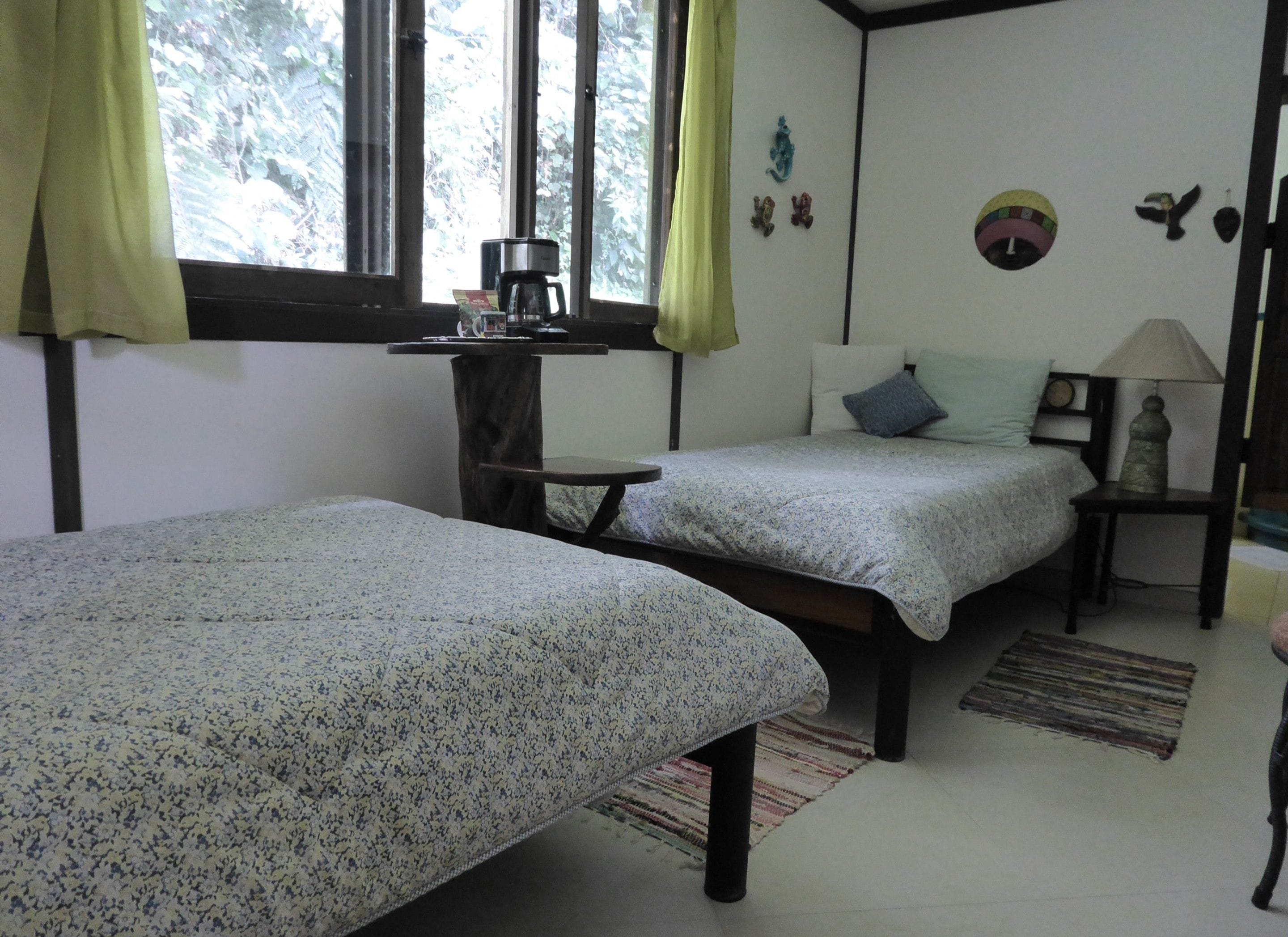 A room with two single beds, a small table with a coffee maker, a lamp, and a window with curtains.