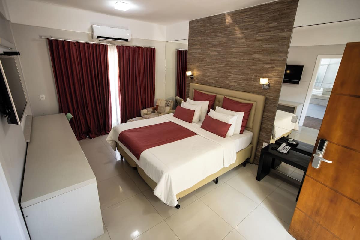 A hotel room with a large bed, a desk, a chair, a TV, and an air conditioning unit.