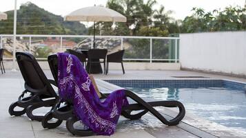 Área à beira da piscina com espreguiçadeiras e uma mesa, rodeada por um muro branco e vegetação ao fundo.