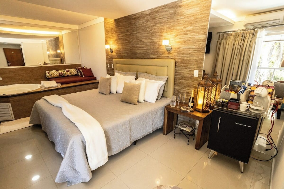 A modern hotel room with a large bed, a bedside table with a lamp, a minibar, and a view of the outdoors.