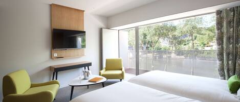 A modern hotel room with two beds, a flat-screen TV, a small table with a glass, and a view of greenery through large windows.
