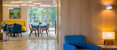 A modern hotel lobby with a blue armchair, a wooden wall, and a dining area with orange chairs.