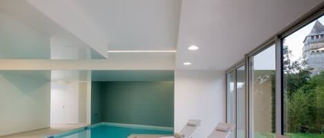 A modern indoor swimming pool with clear glass walls, a tiled wall in the background, and lounge chairs with towels.