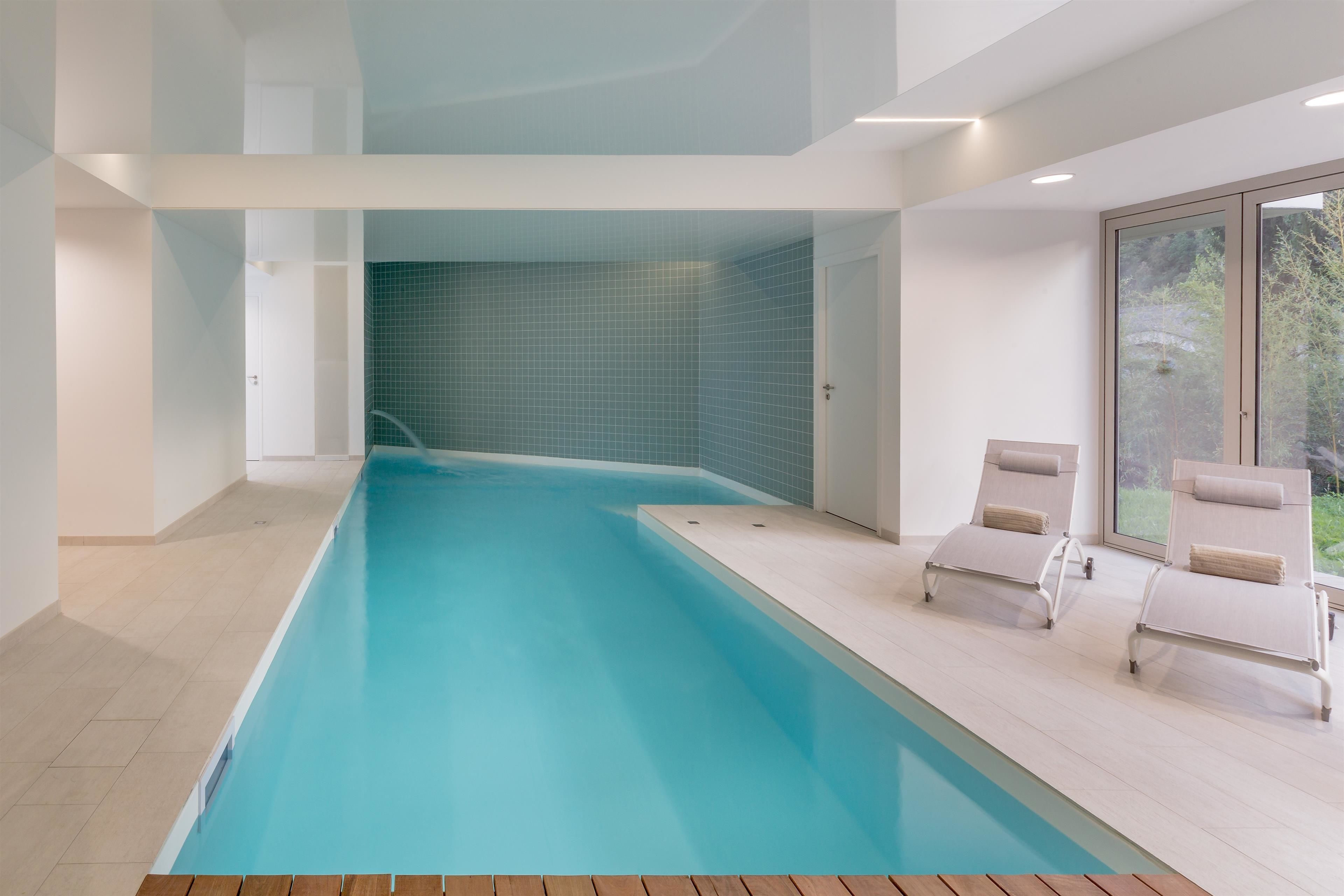 Une piscine intérieure moderne, avec une eau d’un bleu cristallin, un mur carrelé et deux chaises longues placées au bord du bassin.