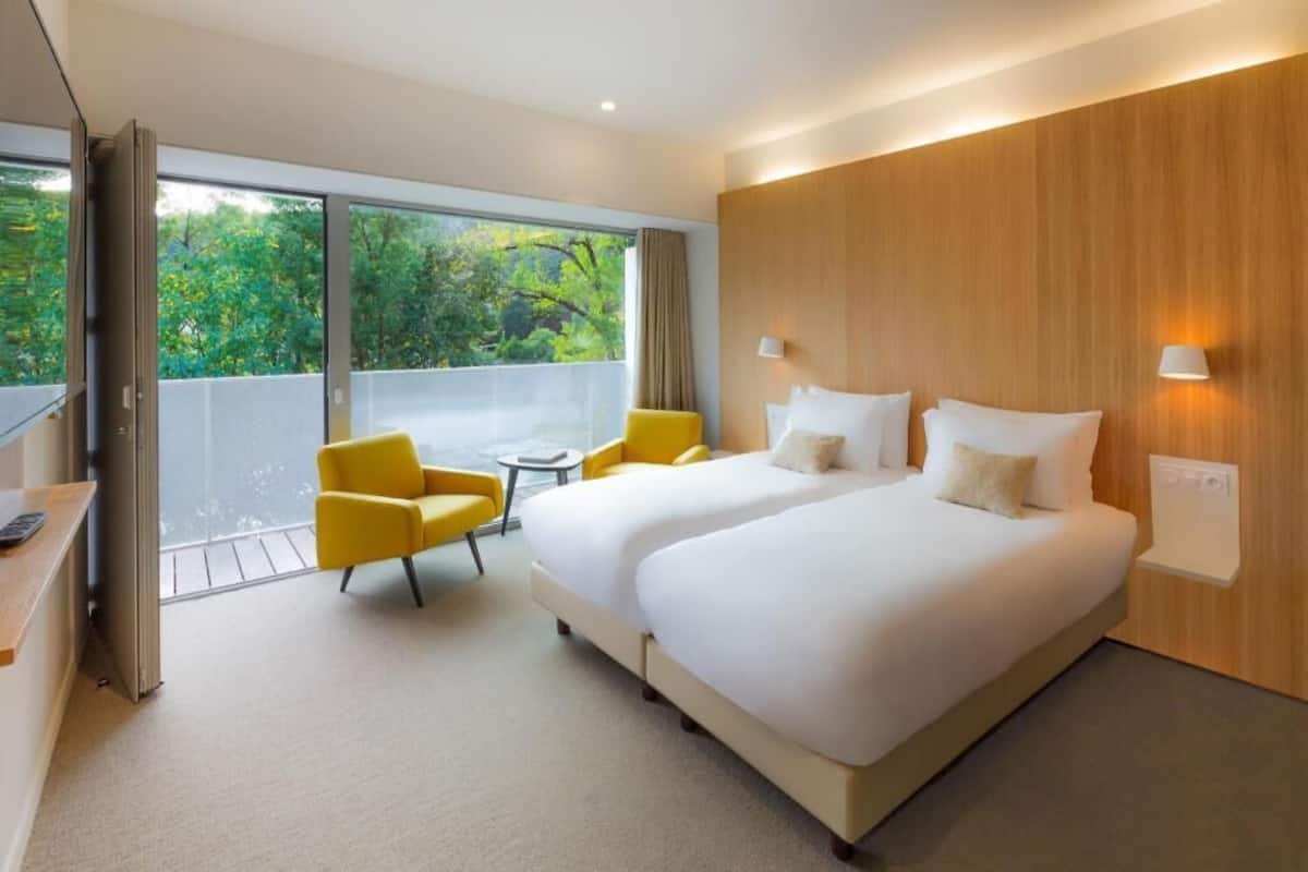 Une chambre d’hôtel avec deux lits, un bureau, une chaise et une vue sur la verdure grâce à de grandes fenêtres.