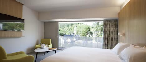 Una habitación de hotel moderna con una cama grande, un sillón amarillo, una mesita redonda y un ventanal que ofrece vistas a zonas verdes y un puente.