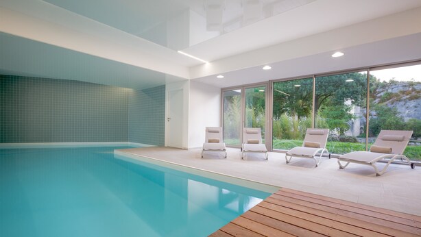 Ein modernes Hallenbad mit großen Glaswänden, Holzterrasse und Liegestühlen.