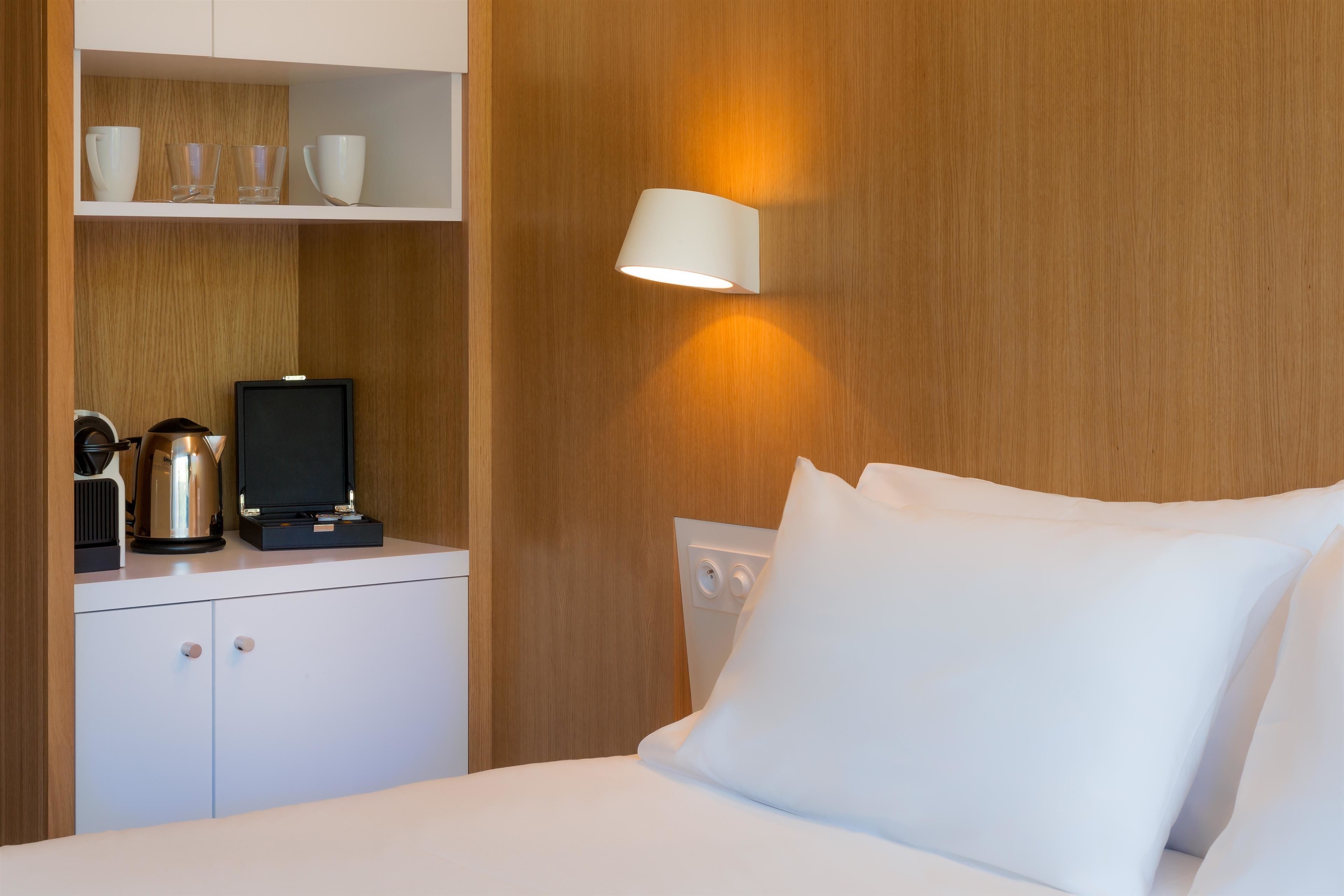 A hotel room with a bed, white bedding, a nightstand with a kettle, cups, and a television.