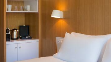 A hotel room with a bed, white bedding, a nightstand with a kettle, cups, and a television.