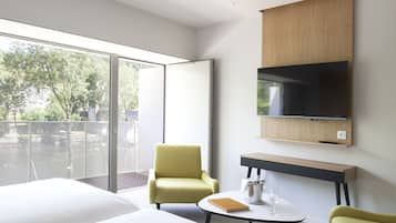 Ein modernes Hotelzimmer mit einem großen Bett, einem grünen Sessel, einem kleinen runden Tisch mit einem Weinglas und einem Buch, einem an der Wand montierten Fernseher und einem Balkon mit Blick auf Bäume.