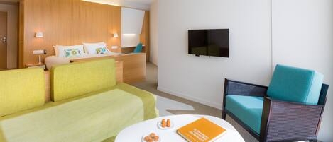 A hotel room with a bed, a green sofa, a small table with a book and snacks, and a TV on the wall.