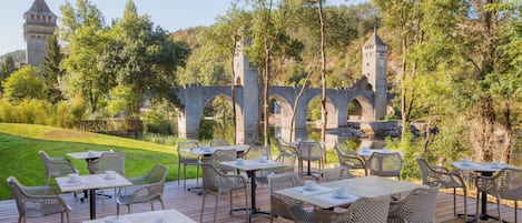Un espacio de comedor al aire libre con mesas y sillas, con vistas a un puente histórico y una torre.