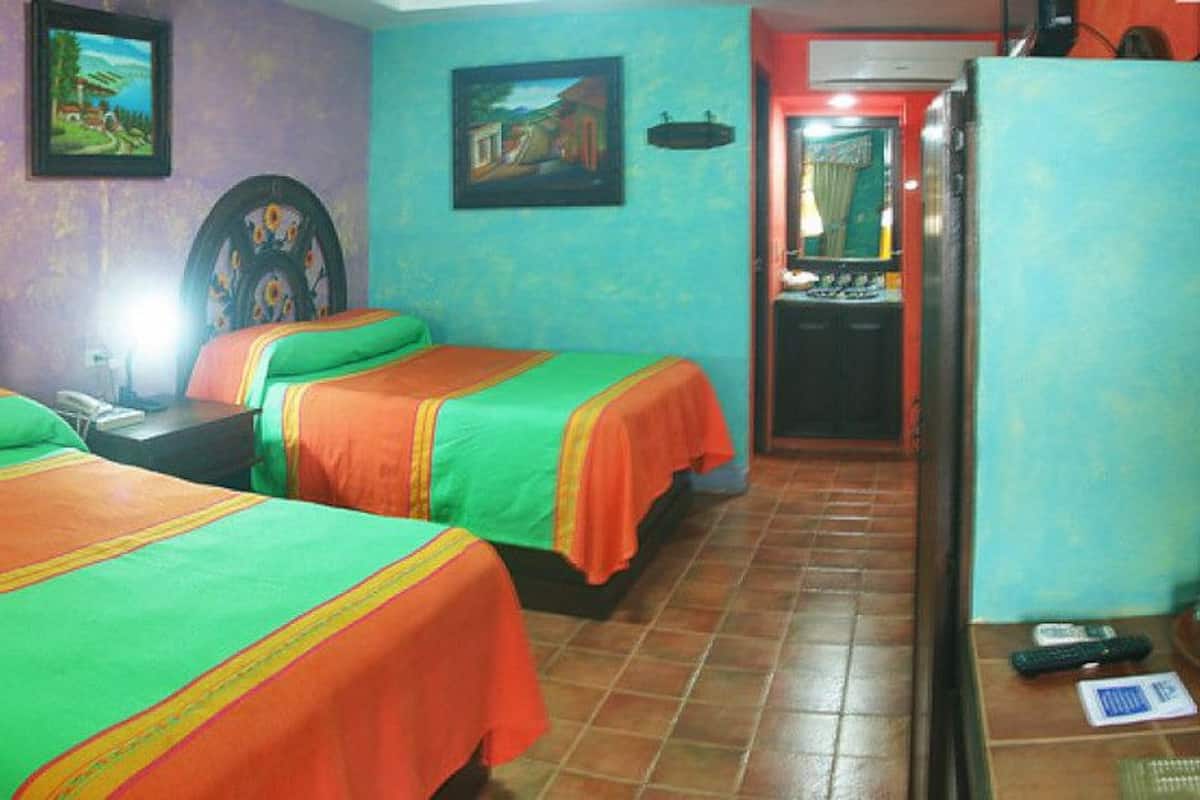 A room with two beds, each with a floral headboard, a small table, and a chair. There is a door leading to a kitchen area.