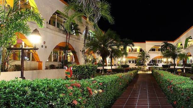 Un patio de hotel por la noche con un camino central, palmeras y edificios bien iluminados.