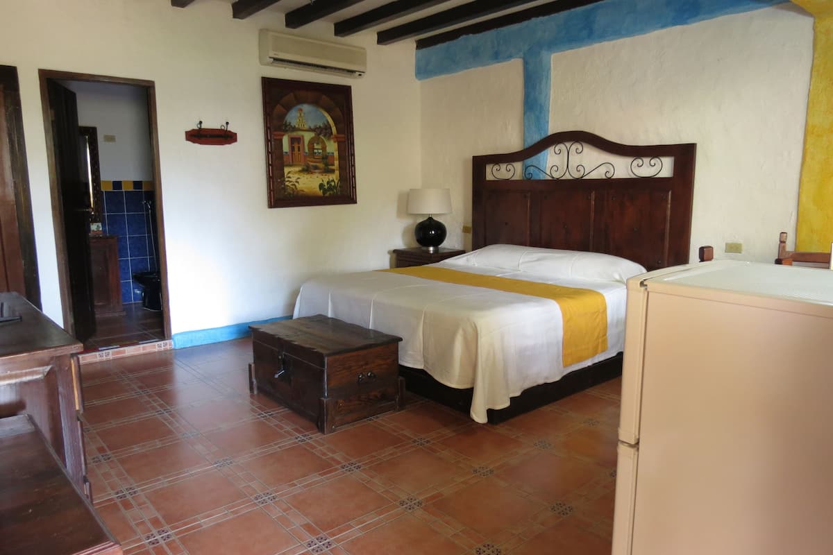 A bedroom with a bed, a wooden chest, a nightstand with a lamp, and a wall-mounted air conditioner.