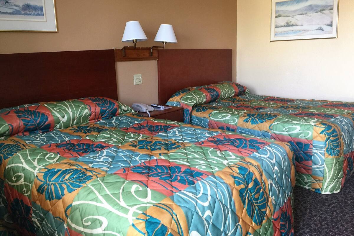 A hotel room with two beds, each with a colorful floral bedspread, a wooden headboard, two bedside lamps, a telephone, and a painting on the wall.