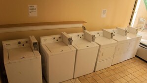 A laundry room with multiple Whirlpool washers and dryers, a wooden shelf above, and a tiled floor. - Bama Inn (Huntsville)
