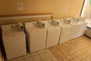 A laundry room with multiple Whirlpool washers and dryers, a wooden shelf above, and a tiled floor.