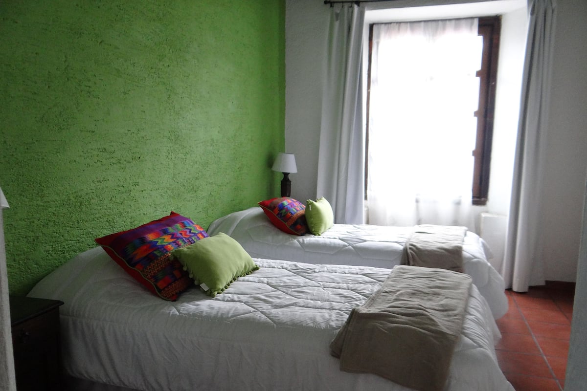 Una habitación con dos camas, paredes verdes, una ventana con cortinas y una lámpara de noche.
