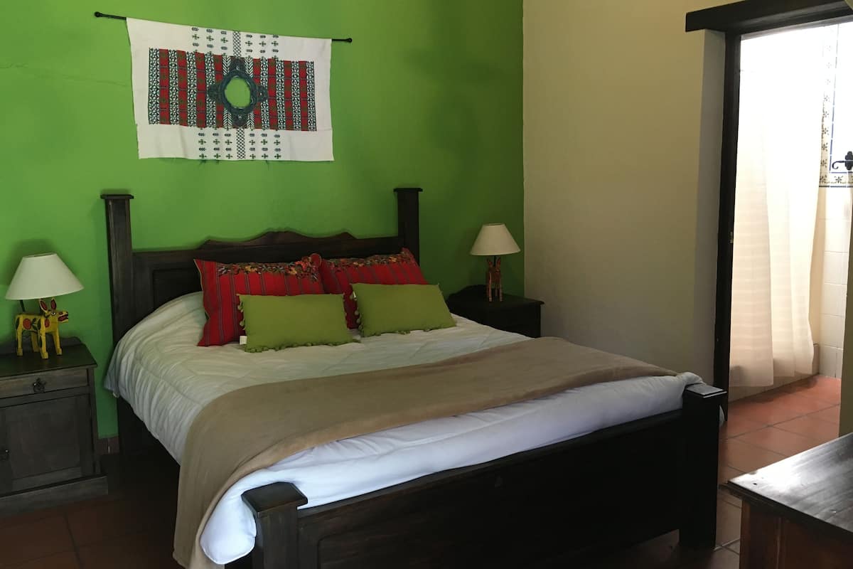Un dormitorio con cama, paredes verdes, cabecero de madera, dos mesitas de noche con lámparas y una ventana con cortinas.