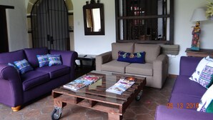 A living room with a purple sofa, a beige sofa, and a glass coffee table with magazines. There is a large window with bars and a decorative mirror above it. - Hotel Casa Naranjo (Antigua Guatemala)