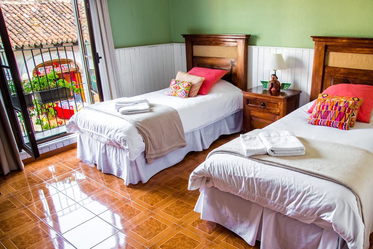 Een hotelkamer met twee bedden, een houten hoofdbord, een nachtkastje en uitzicht op een balkon met planten.