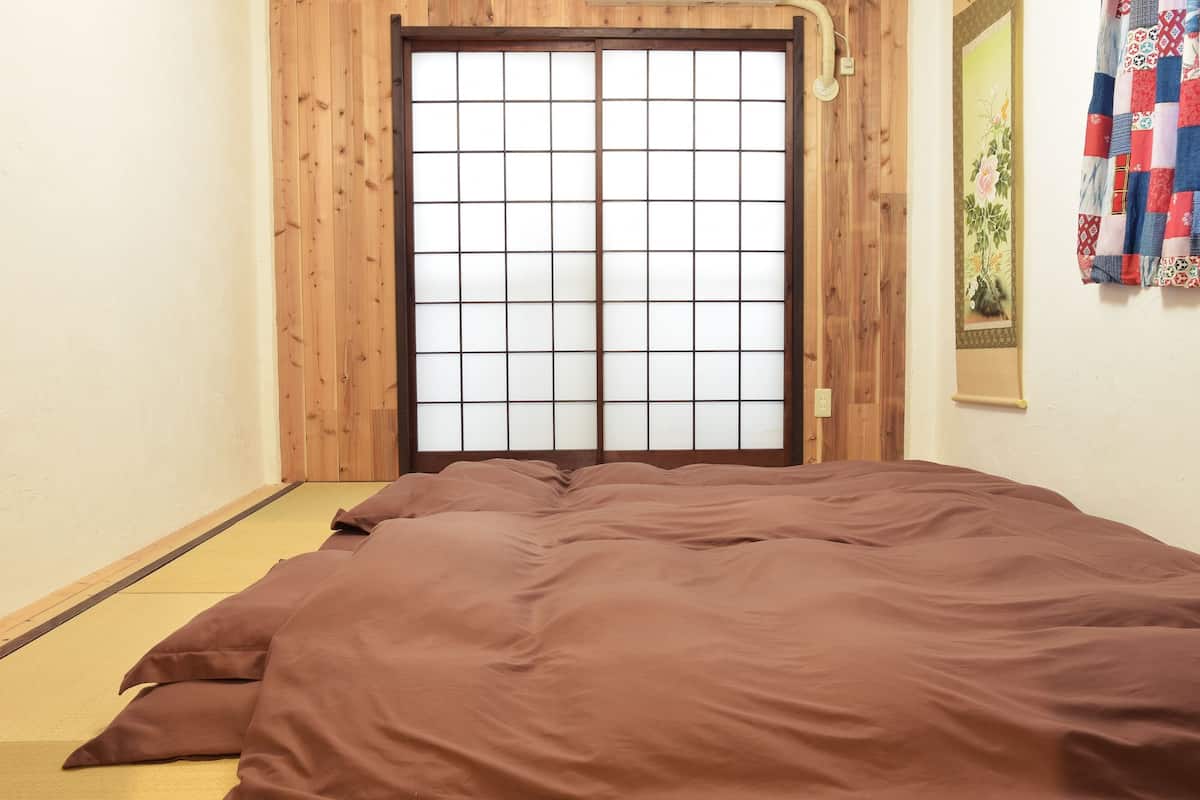 Una habitación con suelo de tatami, puerta de shoji de madera y una cama con manta marrón.