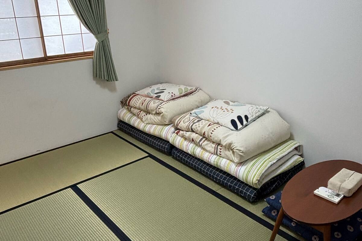A traditional Japanese-style room with tatami flooring, a futon bed, a small round table, and a window with a curtain.