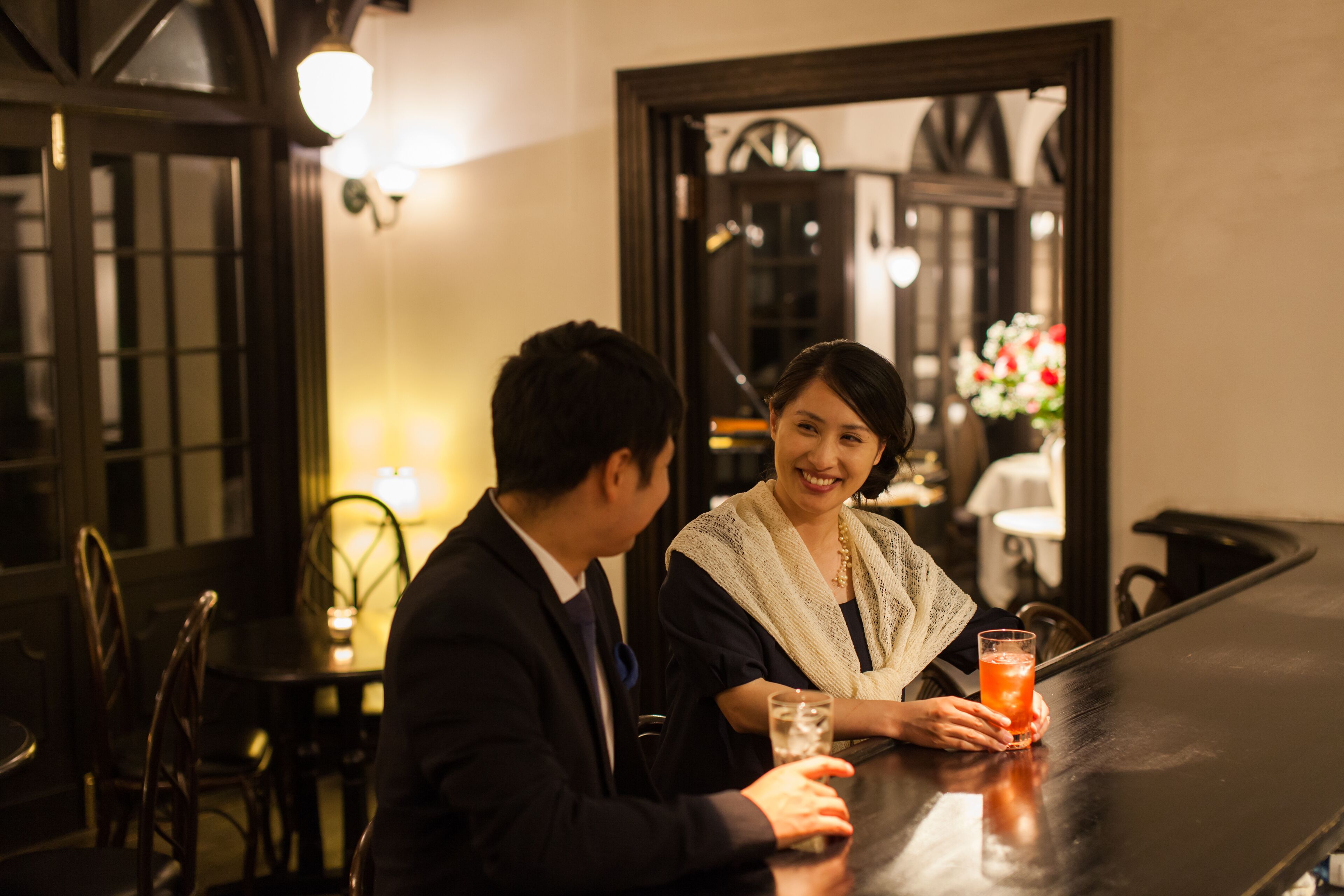 A man and a woman seated at a bar in a dimly lit room with wooden furniture and a clock on the wall.
