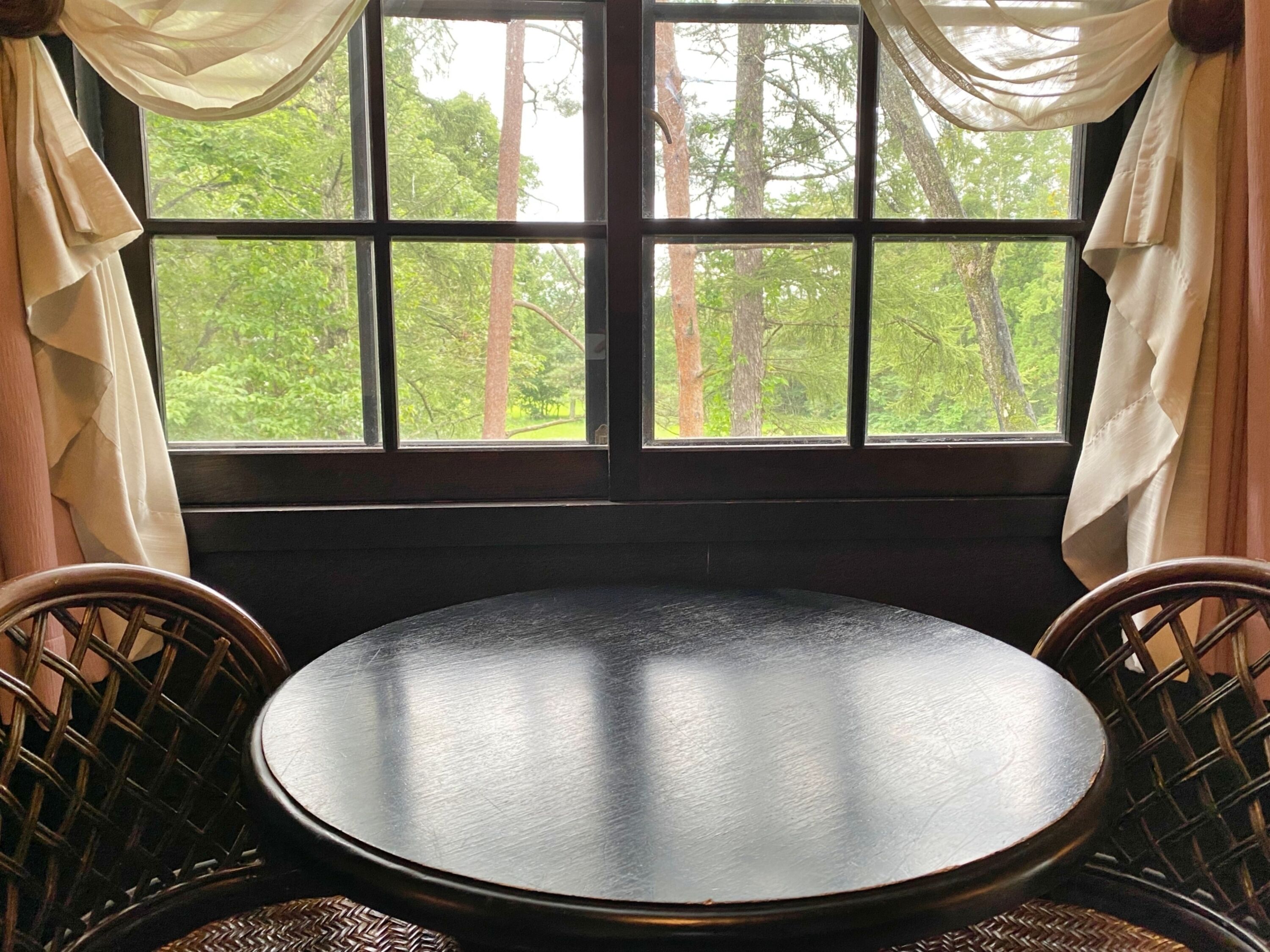 A round table with two wicker chairs in front of a window with multiple panes, curtains, and a view of trees.