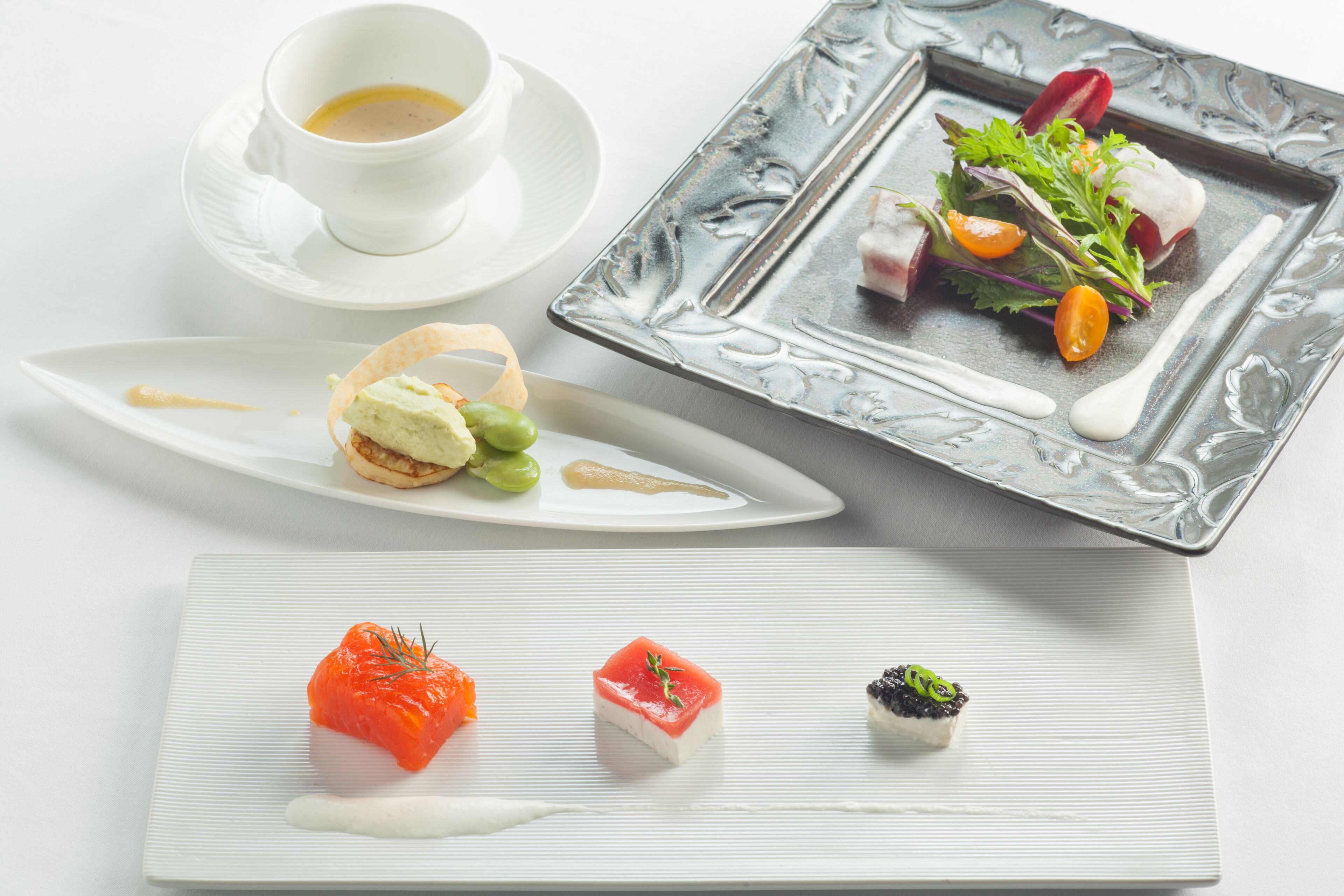 A variety of plated dishes, including a salmon tartare, a small tart, and a sushi roll, arranged on a white table.