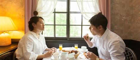 Una mesa de comedor con alimentos para el desayuno, una lámpara y una ventana con cortinas.