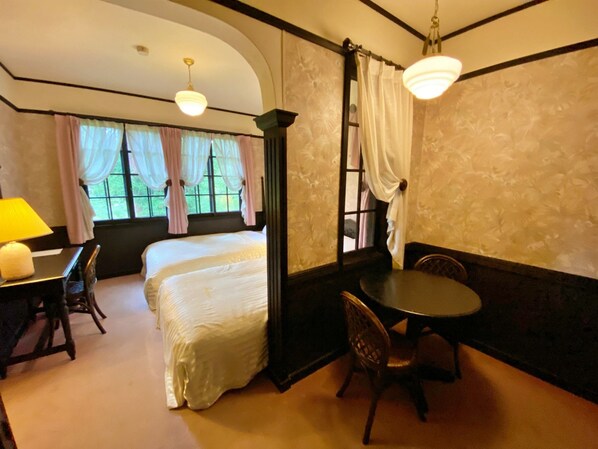 Habitación de hotel con dos camas, una mesa de comedor y sillas. Hay cortinas en las ventanas y un candelabro cuelga del techo.
