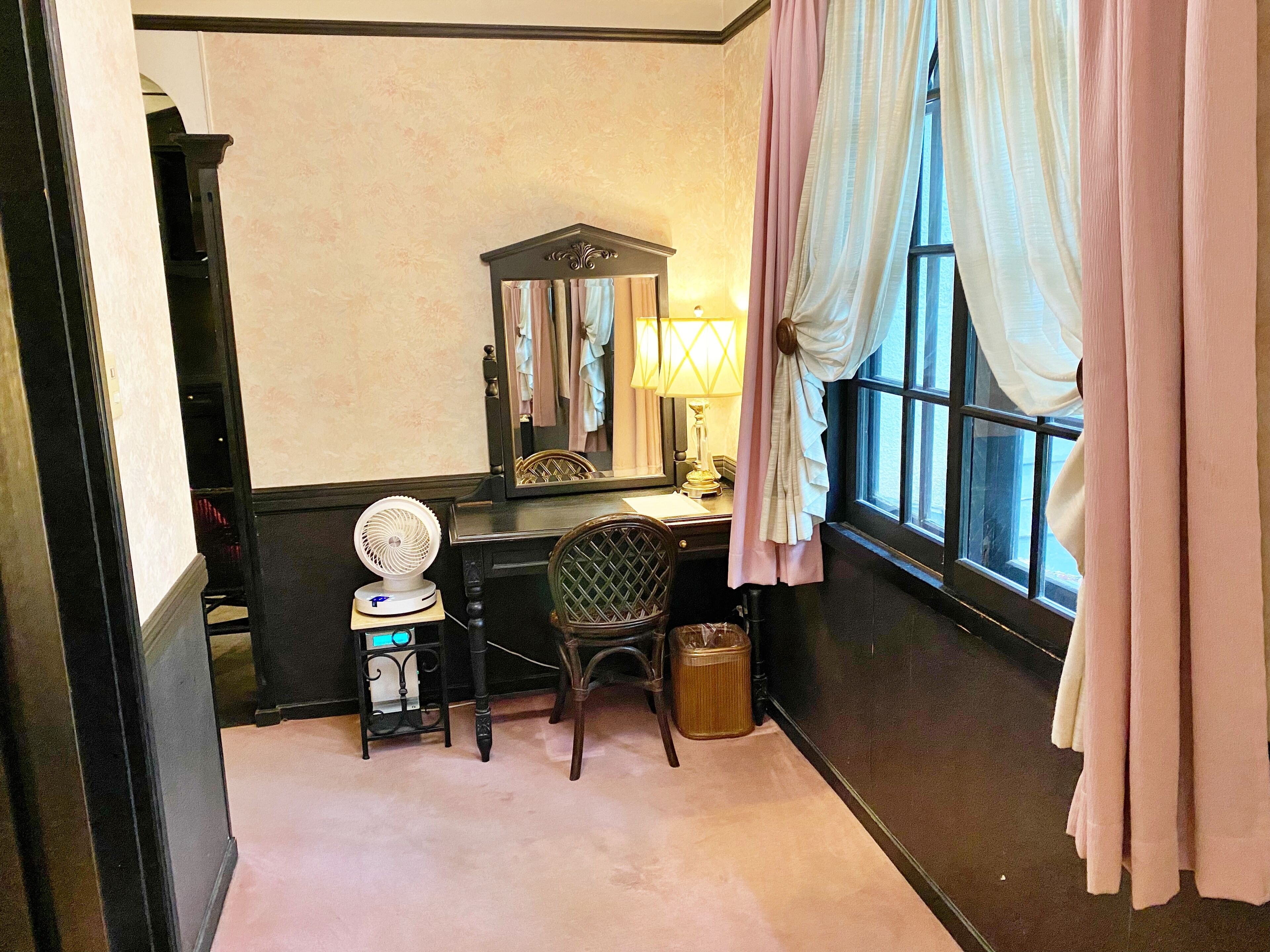 A room with a desk, chair, mirror, fan, and window with curtains.