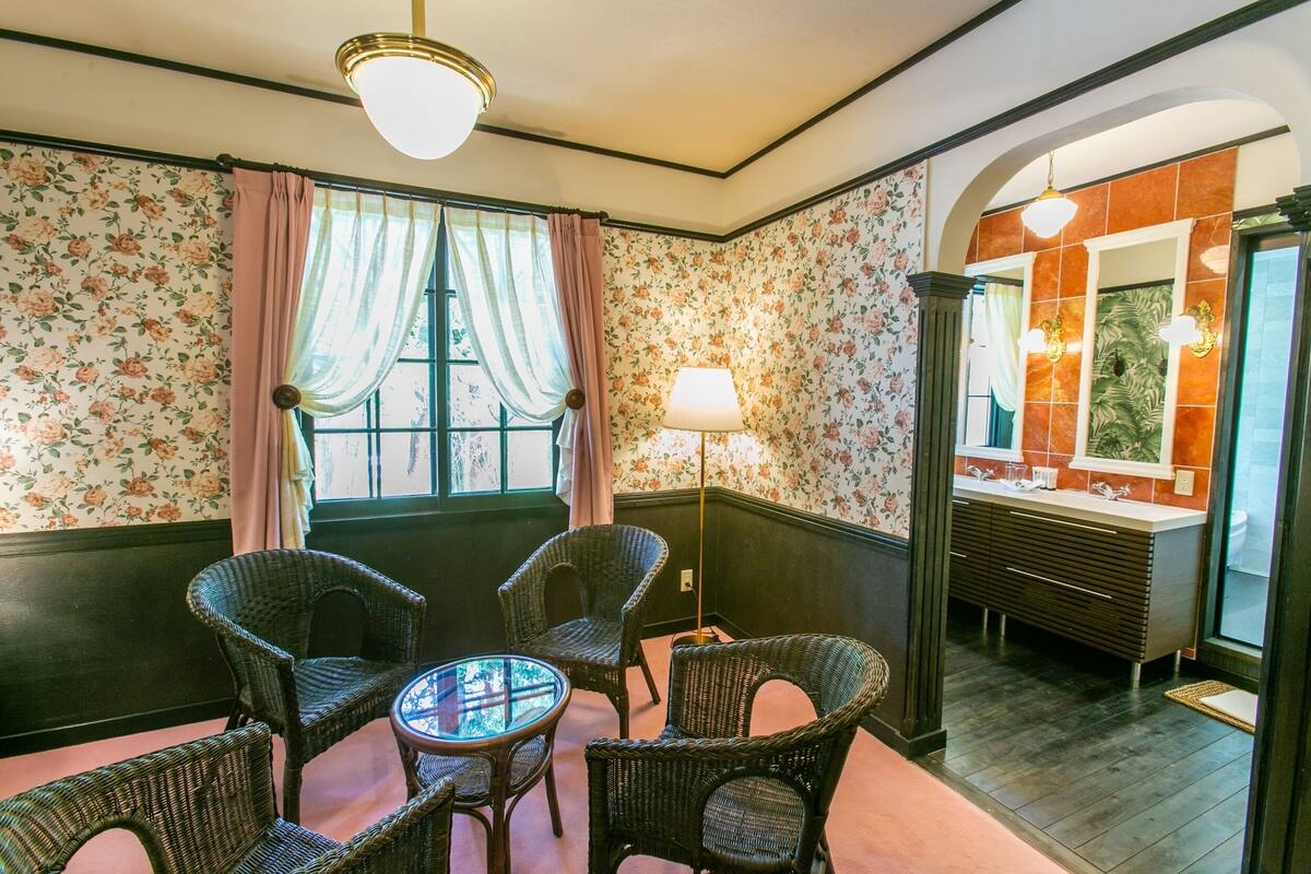 A room with wicker chairs, a glass table, floral wallpaper, a window with curtains, a lamp, and a bathroom with a mirror and sink.