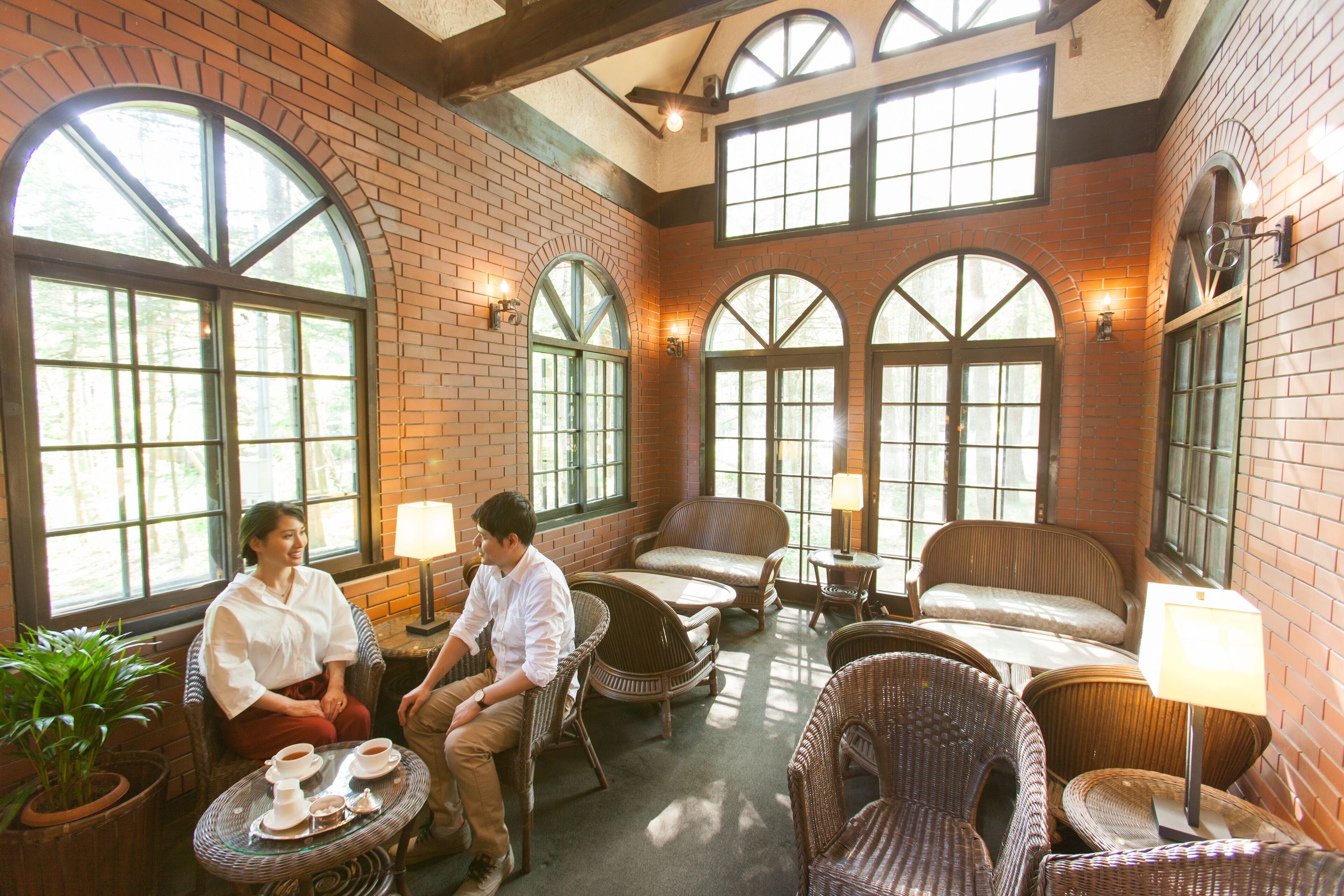 A cozy indoor seating area with wicker furniture, large windows, and a brick wall.