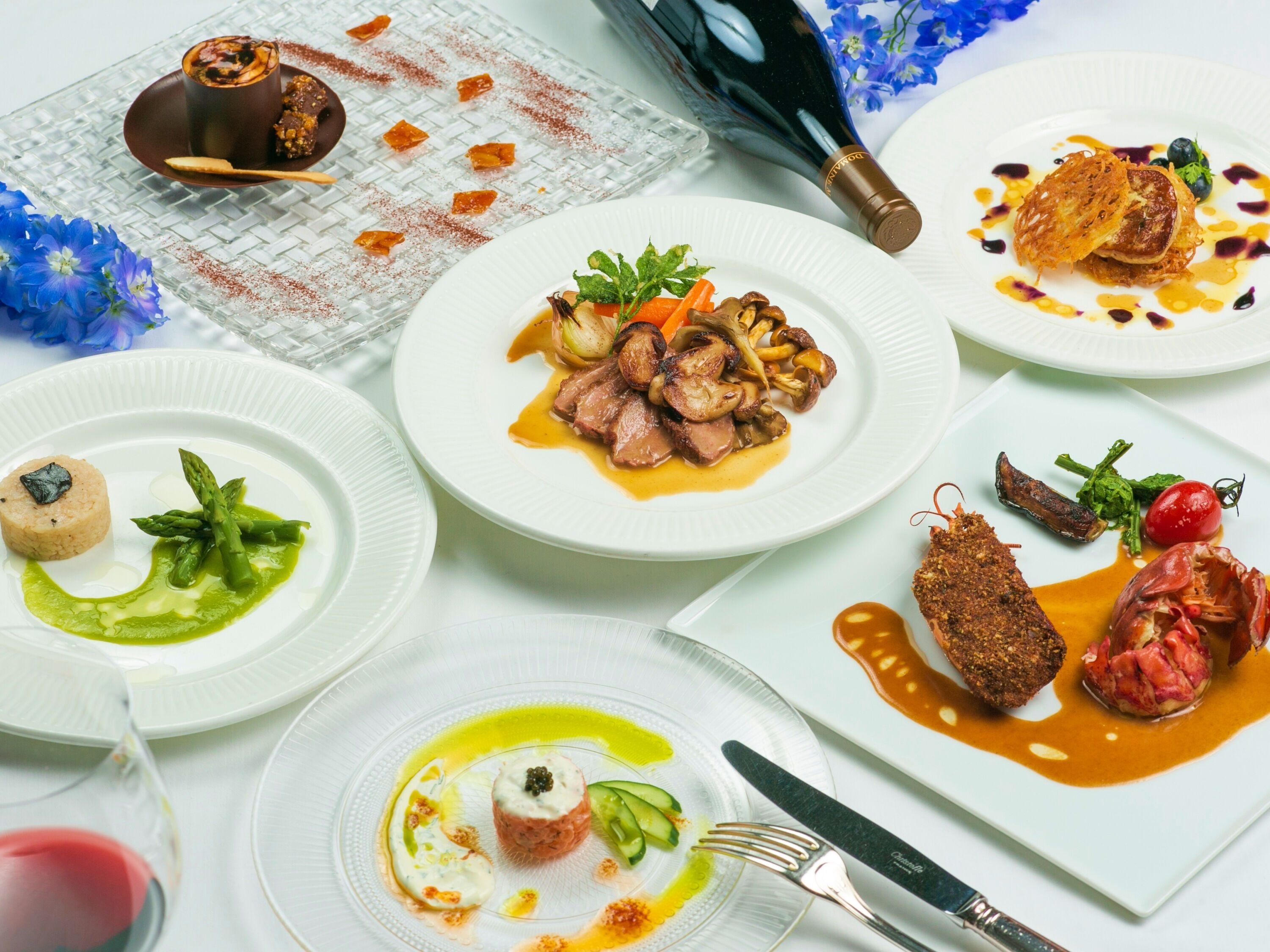 A variety of dishes on white plates, a wine bottle, and a glass of wine.