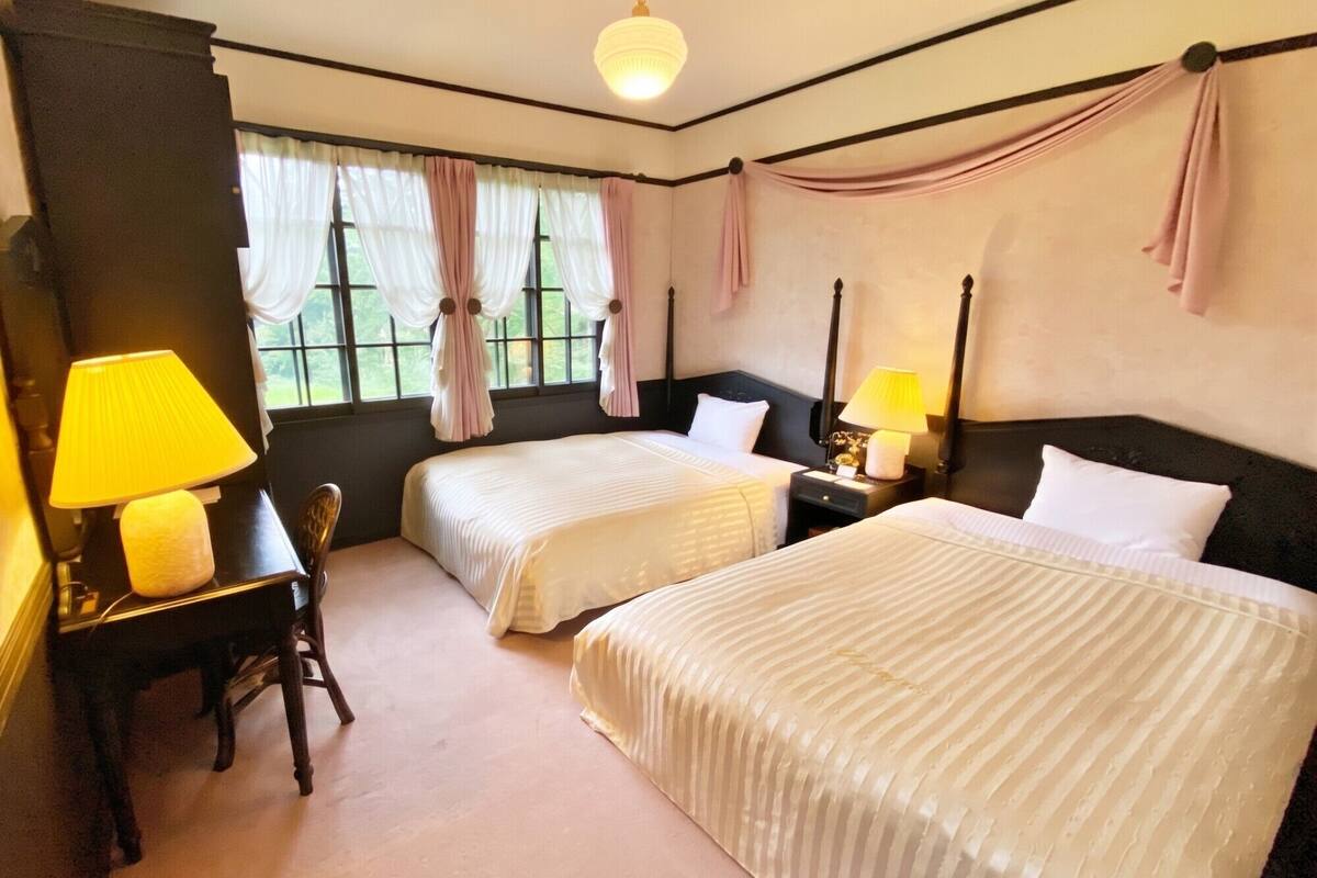 A hotel room with two beds, a desk, a chair, a lamp, and a window with curtains.