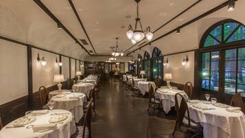 A long dining hall with tables set for service, large windows, and chandeliers.