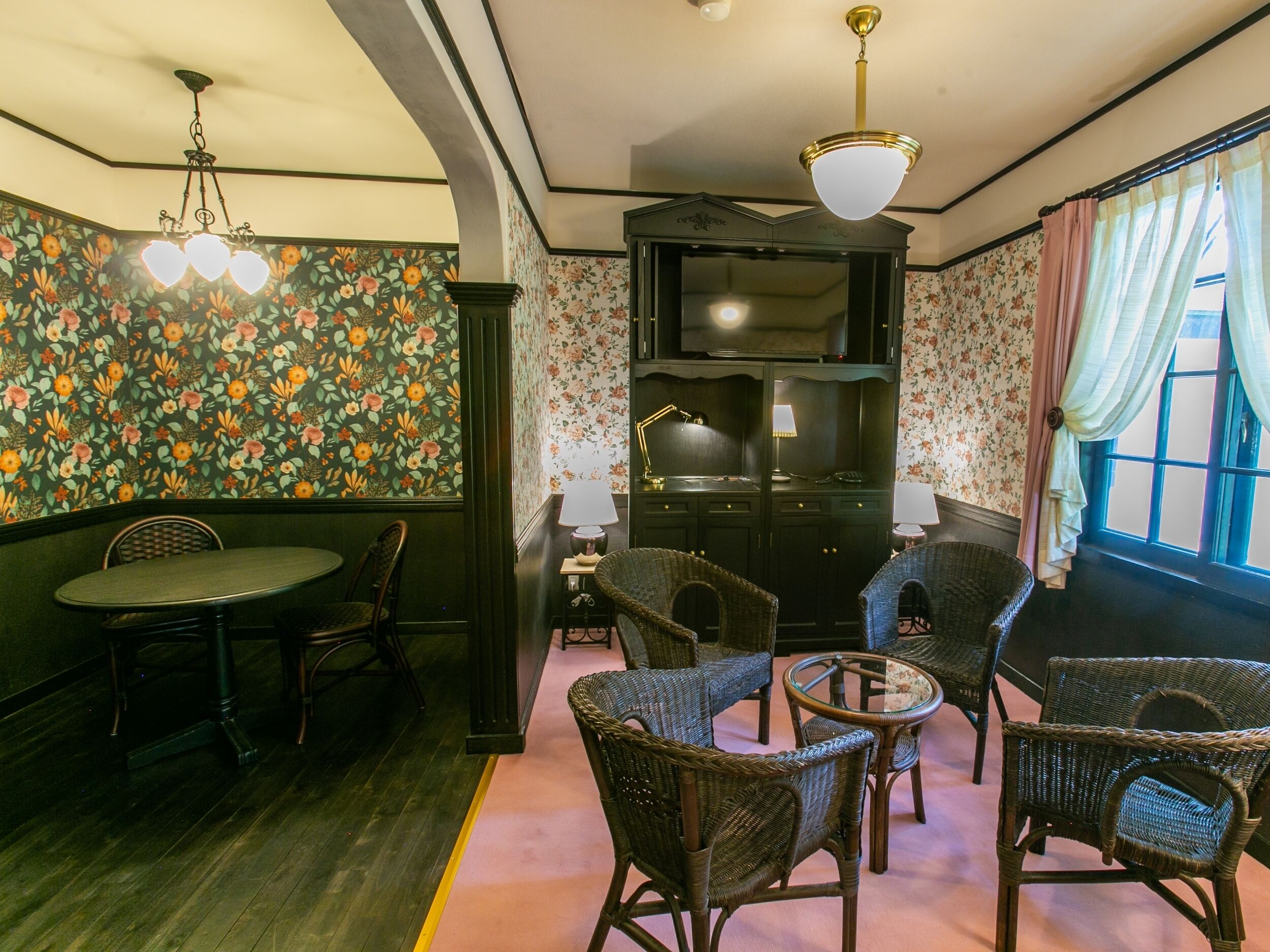 Una habitación con una mesa de comedor y sillas, televisor, muebles de mimbre y papel tapiz con motivos florales.