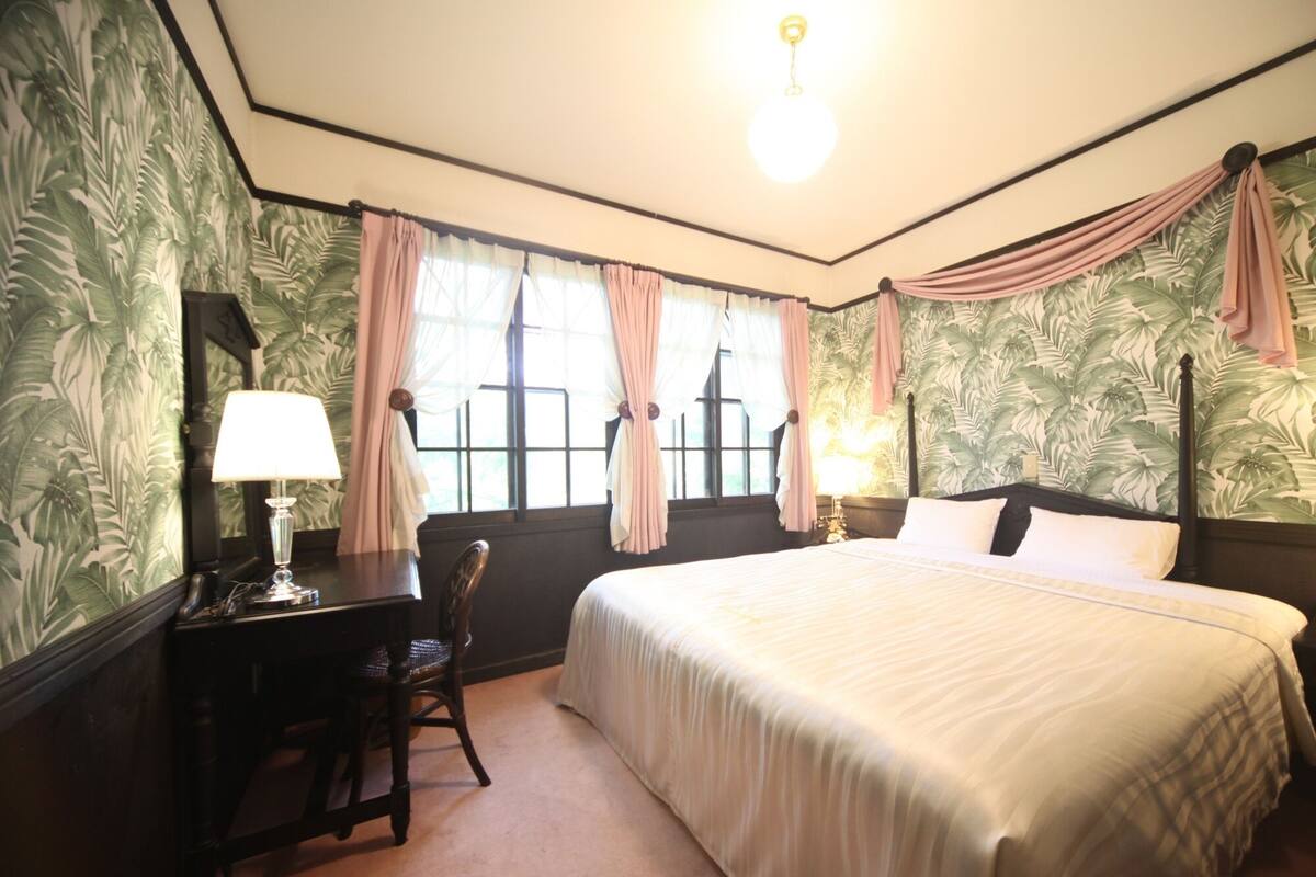 A bedroom with a large bed, a desk with a lamp, and a chair. There are curtains on the windows, and the walls have a leafy pattern.