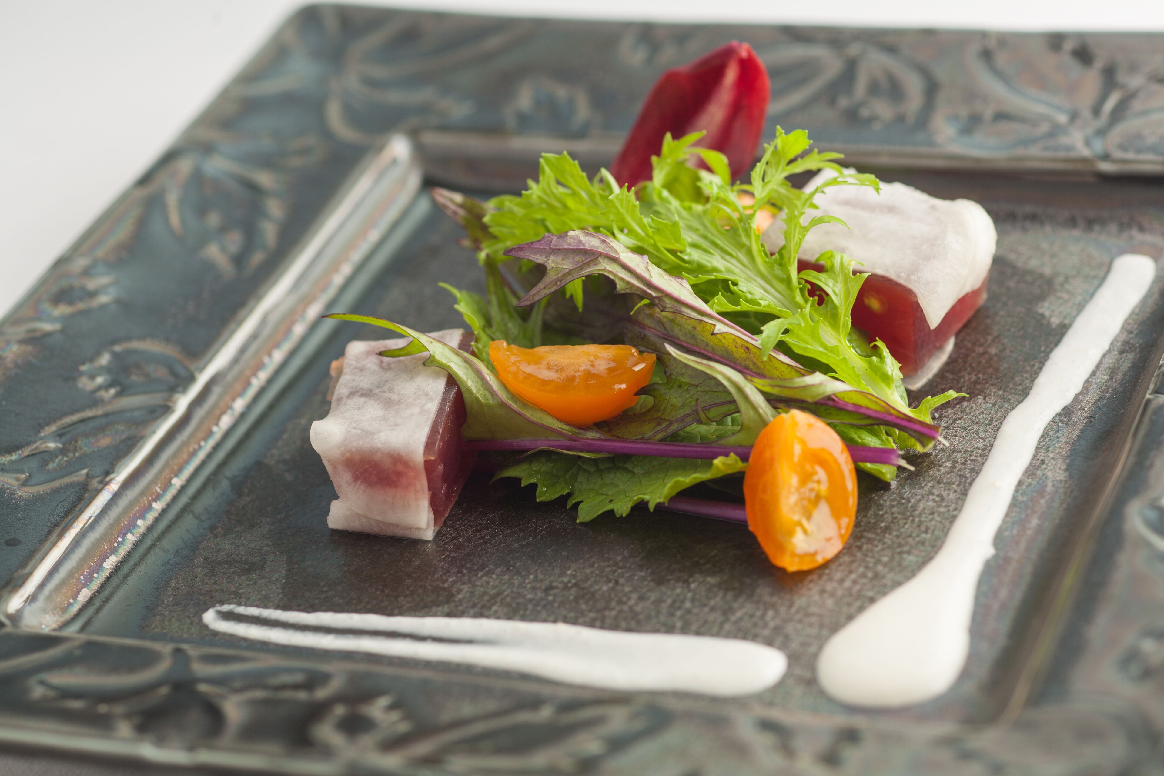 Une assiette carrée ornée d’un motif décoratif, contenant une salade composée de légumes verts, de tomates cerises et d’un morceau de viande, accompagnée d’une cuillère blanche.