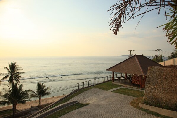 A beachside view with a covered seating area, palm trees, and a clear view of the ocean. - Lima Satu Resort (Pemenang)
