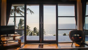 A room with a large sliding glass door leading to a balcony with a view of the ocean and palm trees. - Lima Satu Resort (Pemenang)